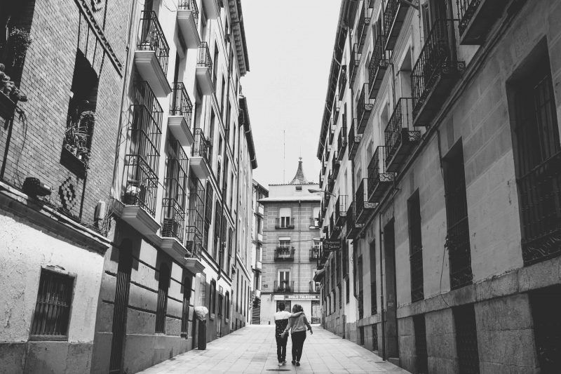 Plano general de una calle con edificios a los lados, por cuyo centro camina una pareja de espaldas. La mujer está tocando con su mano el culo del hombre